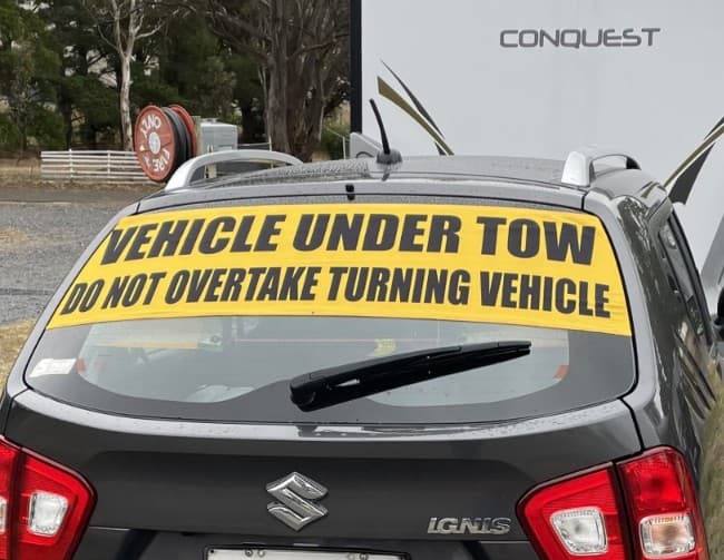 Tow Smart Regular sign on the outside of a Suzuki Ignis rear window: Vehicle Under Tow, Do Not Overtake Turning Vehicle